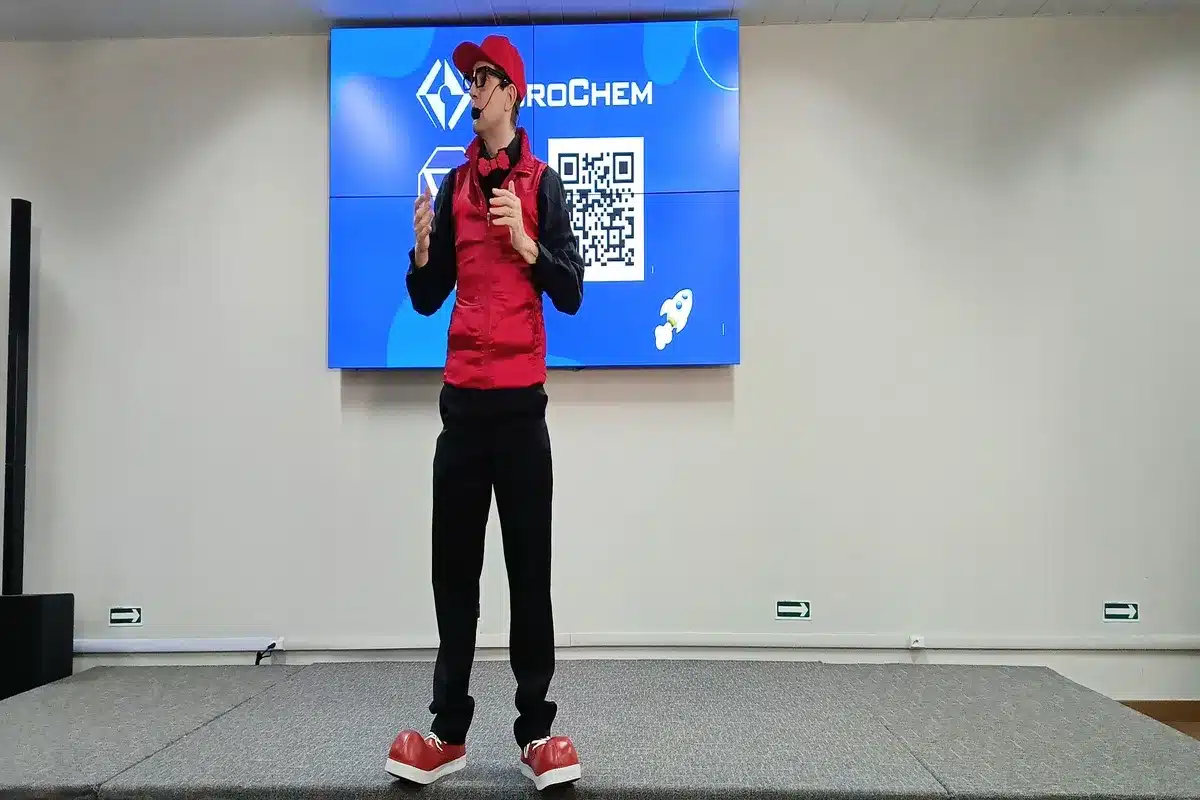 Homem fantasiado com colete vermelho, boné e sapatos grandes, fazendo uma apresentação em um palco em frente a um telão com o logo da EuroChem e um código QR.
