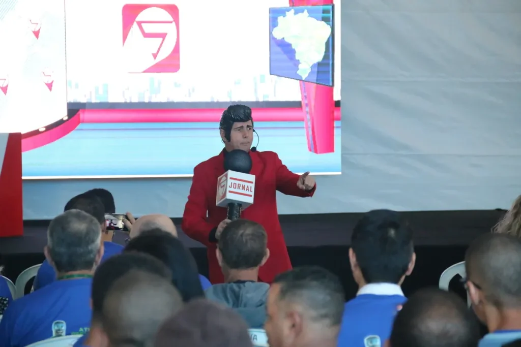 Palestrante vestido como jornalista ou âncora de TV (casaco vermelho e microfone "JORNAL") gesticula no palco com um mapa do Brasil ao fundo, em uma palestra sobre vacinação.