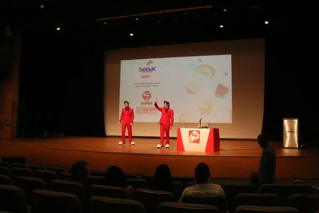 Dois homens vestidos com ternos vermelhos, representando o rock and roll, em um palco, ministrando uma palestra divertida sobre vacinação em um auditório.