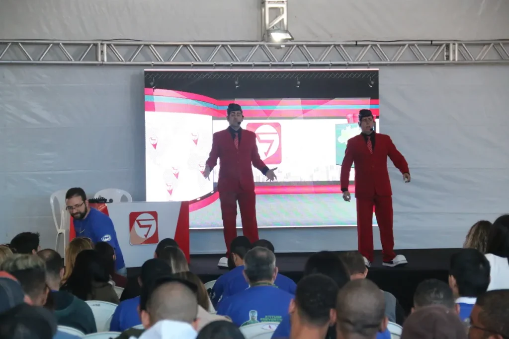 Dois palestrantes idênticos, de terno e gravata vermelhos, gesticulam no palco à frente de um telão digital, em uma palestra sobre vacinação e saúde.
