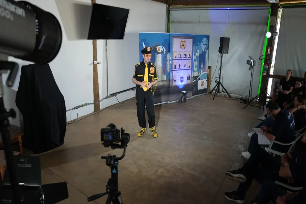 Homem vestido como um policial de trânsito (camisa amarela e preta, boné e óculos escuros) em um estúdio ou galpão com equipamentos de gravação, apresentando para um pequeno grupo, ideal para palestras sobre violência psicológica e assédio moral no trabalho.